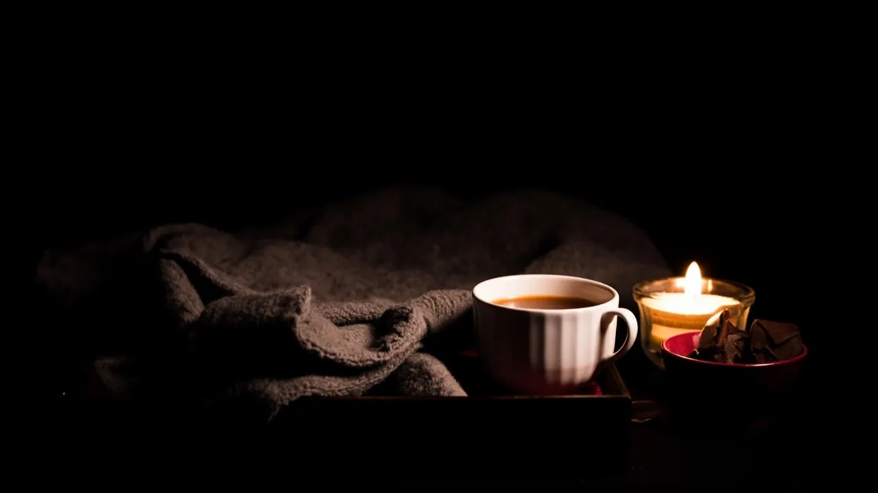 A folded grey blanket, a cup of tea and a small bowl of chocolates on a dark wooden tray by candlelight