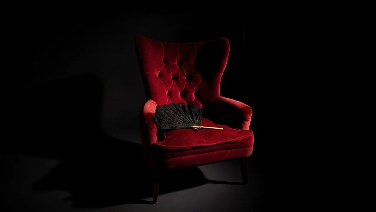A wide red velvet wingback armchair with a black lace fan resting on the seat under a spotlight