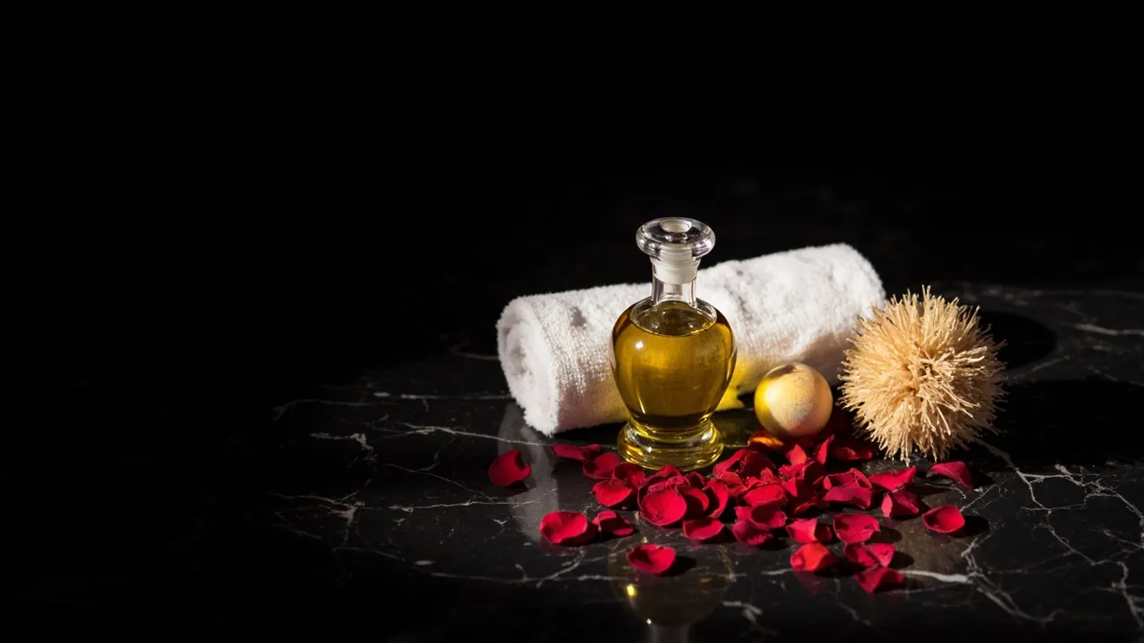 A crystal carafe of golden oil, a rolled white towel and red rose petals on black marble
