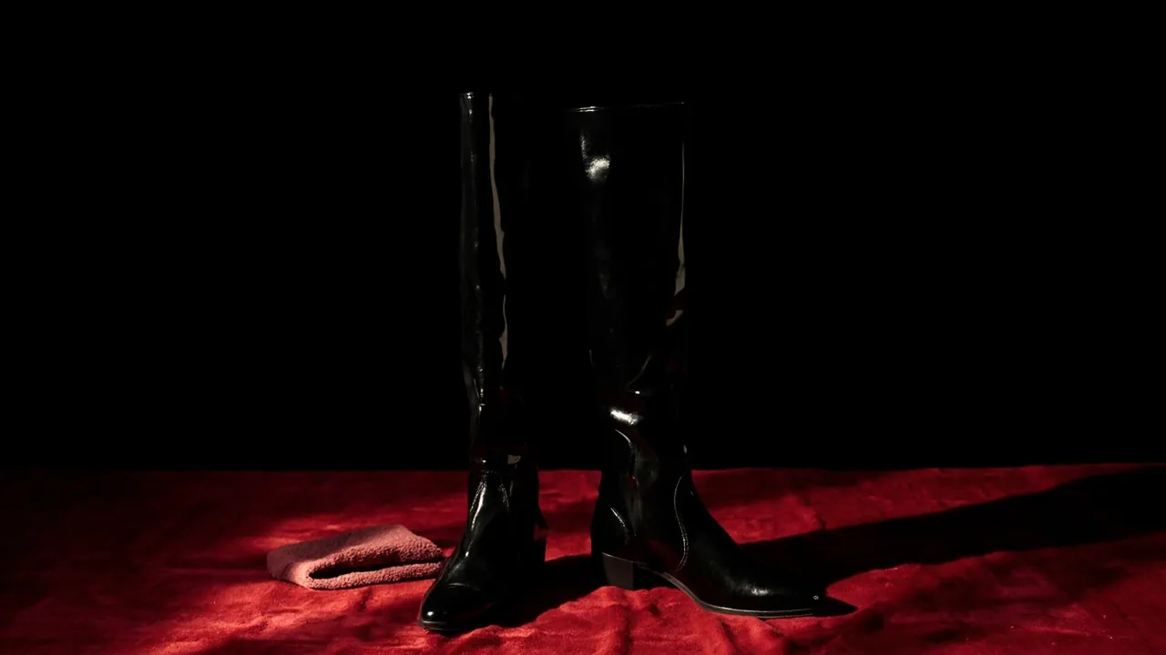 A pair of tall black leather boots standing on crimson velvet beside a soft polishing cloth