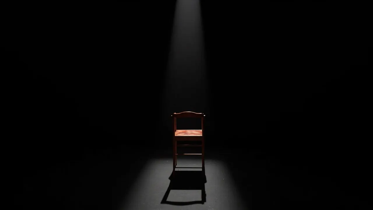 An empty wooden chair facing the corner of a dark room under a single spotlight