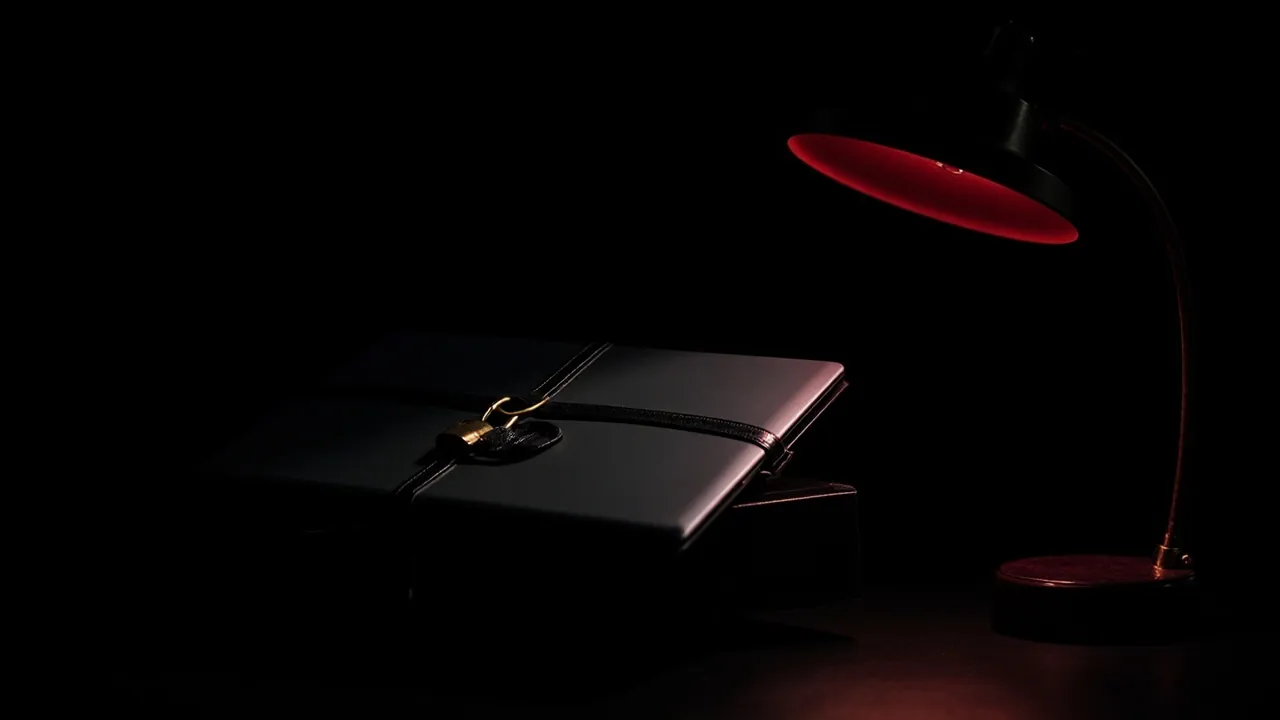 Ein geschlossener schwarzer Laptop, mit einem schwarzen Lederriemen und einem kleinen Messingvorhängeschloss verschlossen, auf einem dunklen Schreibtisch unter einer roten Lampe