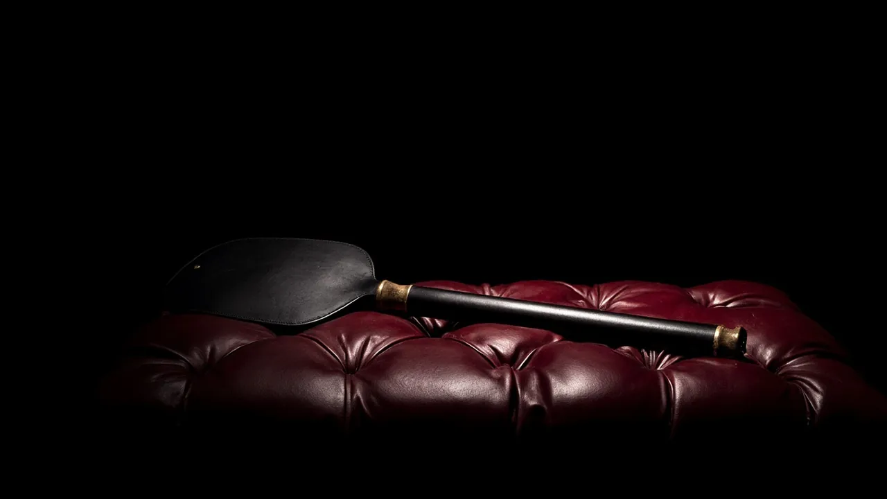 A black leather spanking paddle lying on a tufted burgundy leather bench