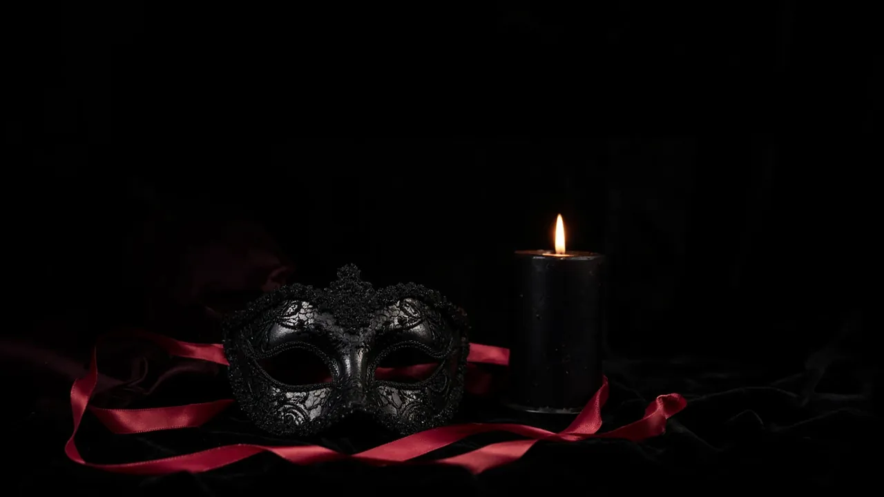 Eine schwarze venezianische Maskenballmaske aus Spitze mit langen roten Satinbändern auf schwarzem Samt neben einer brennenden schwarzen Kerze