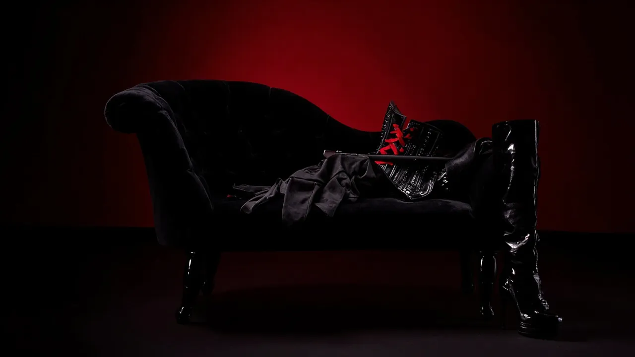A black corset with red lacing, long gloves and a riding crop on an empty velvet chaise, with patent thigh boots beside it