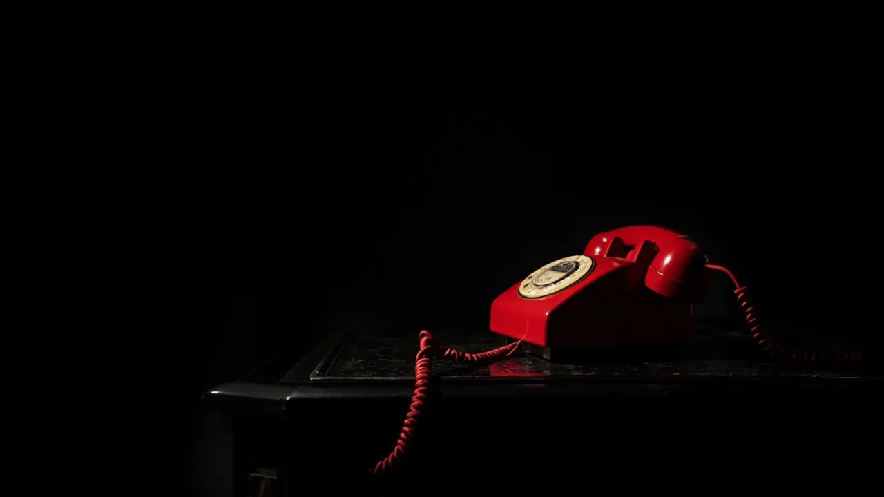 A red rotary telephone on a dark table with its coiled cord trailing over the edge