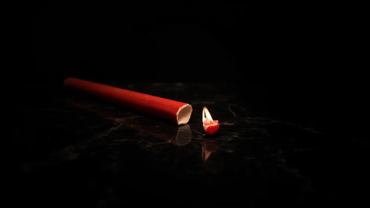 A broken red candle with a small flame at the snapped end on dark marble