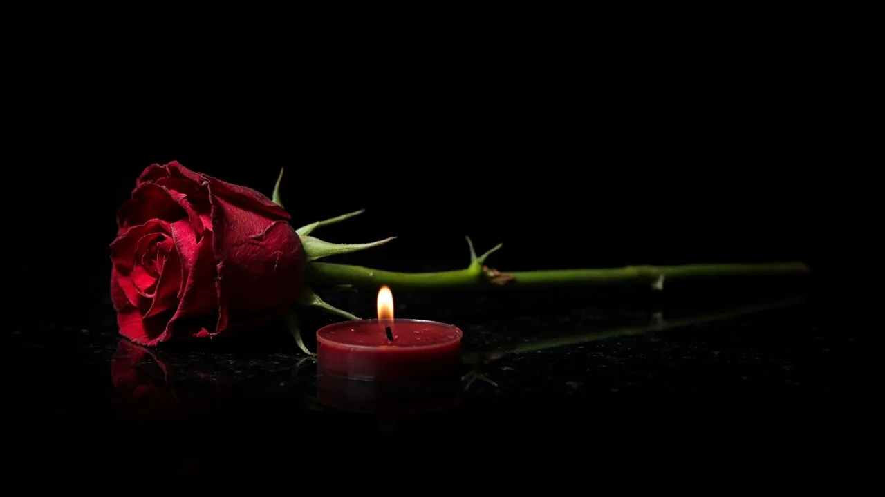 Una rosa rossa a stelo lungo con spine affilate, su marmo nero accanto a una piccola candela rossa