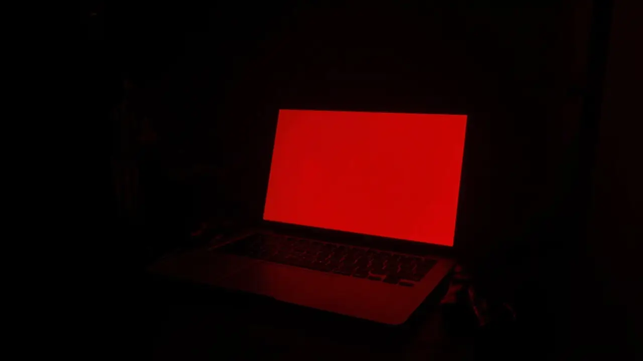 A laptop glowing red in a dark room, black leather gloves beside it